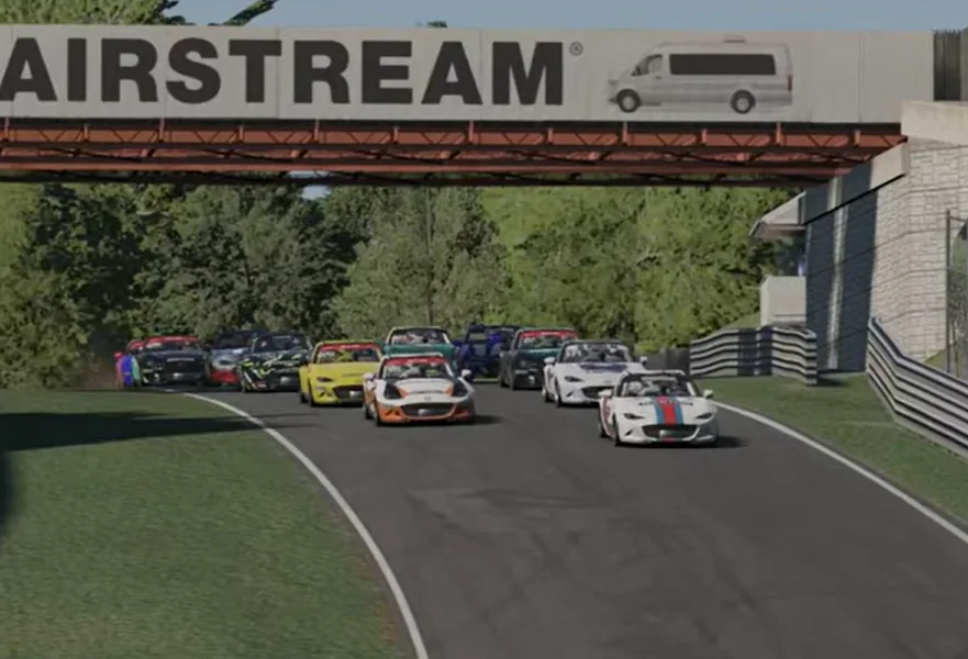 Approaching Airstream Bridge - A full-field view of multiple sports cars driving together on a track as they approach a chicane underneath a large overhead bridge bearing the text 'AIRSTREAM'