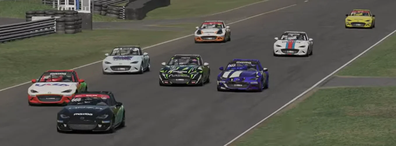 Lead Pack on Straightaway - A high-angle view of a trailing pack of Mazda race cars lined up and accelerating down a straight section of the racetrack next to a grassy border and safety barriers